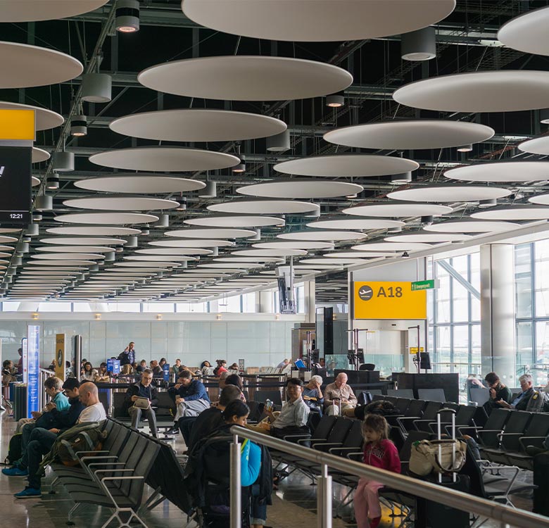 Interior view of a busy airport terminal with travelers seated in waiting areas, modern acoustic ceiling panels, and digital departure signs displaying gate A18 and flight information.