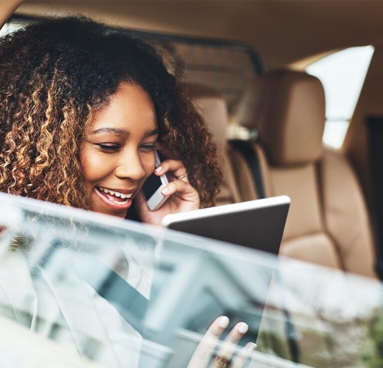 Smiling woman with curly hair talking on a smartphone while holding a tablet in a car. The image depicts a modern and productive lifestyle, highlighting multitasking in a mobile environment.