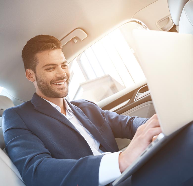 Businessman working on a laptop while sitting inside a modern car, smiling and focused on his tasks, with sunlight streaming through the windows.