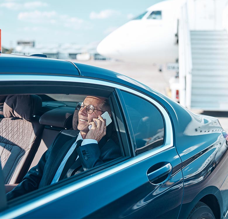 Businessman in suit and glasses sitting in a luxury car at the airport, talking on a smartphone with a plane in the background.