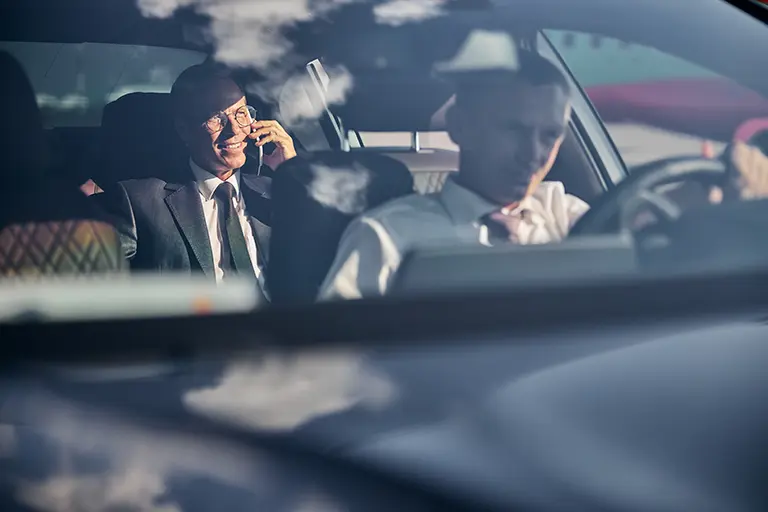 Business professionals engaged in a discussion inside a car, one speaking on a phone and the other focused on driving, showcasing a dynamic work environment.