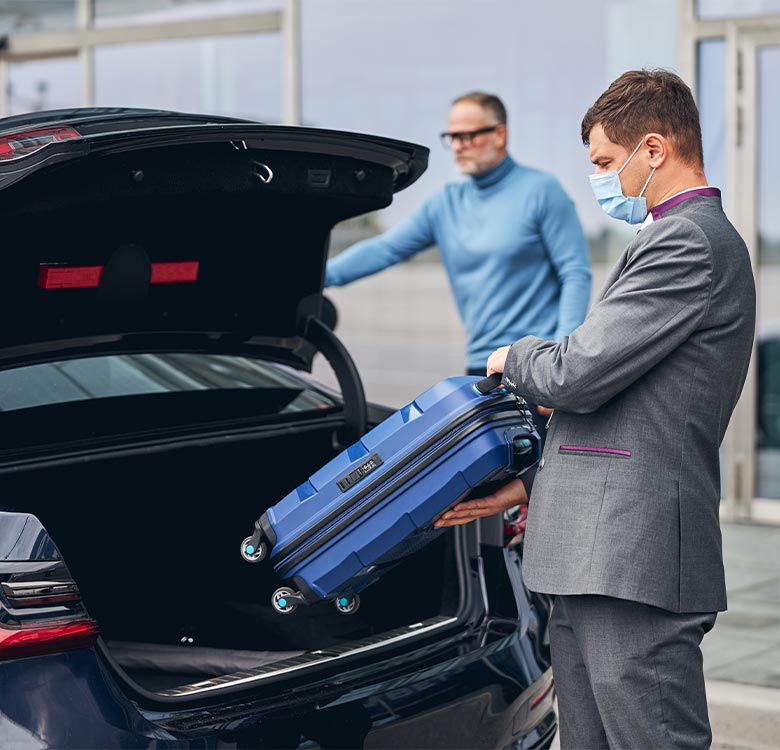 Businessman in a suit loading a blue suitcase into the trunk of a car, with another man in a turtleneck standing in the background, both wearing masks. Ideal for travel-related content or airport transportation services.