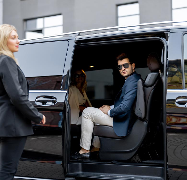 Businessman in sunglasses sitting inside a luxury limousine, while a professional-looking woman stands outside, indicating a high-end transportation service.
