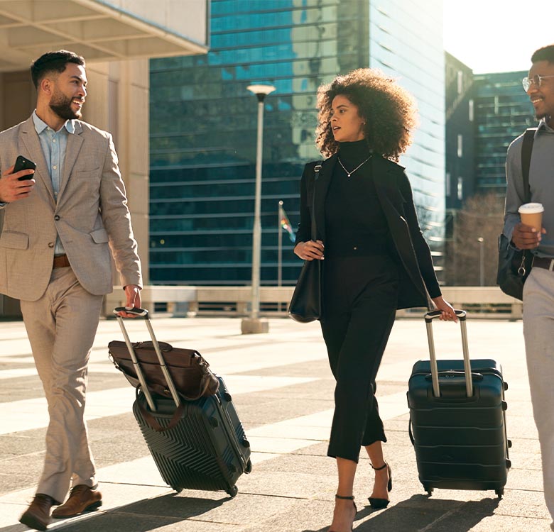 Business professionals walking in a city setting with suitcases, showcasing a blend of modern fashion and urban lifestyle. The scene captures a sunny day with skyscrapers in the background, reflecting a sense of travel and corporate culture.