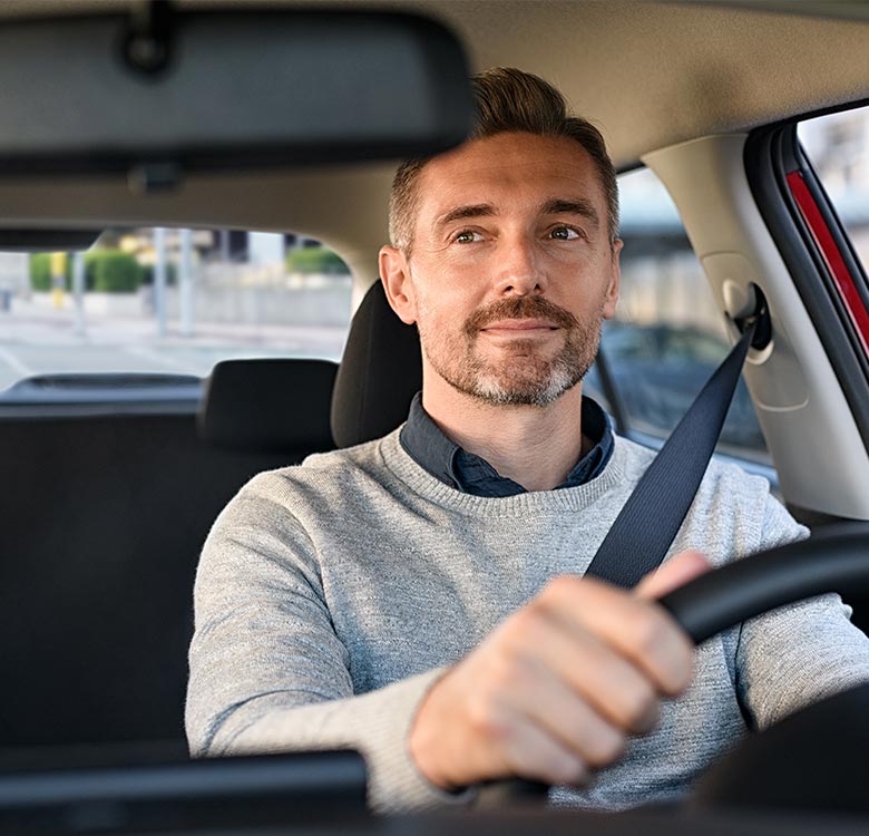 Man driving a car, smiling and looking ahead, wearing a gray sweater and a blue collared shirt, with a seatbelt on, inside a modern vehicle interior.
