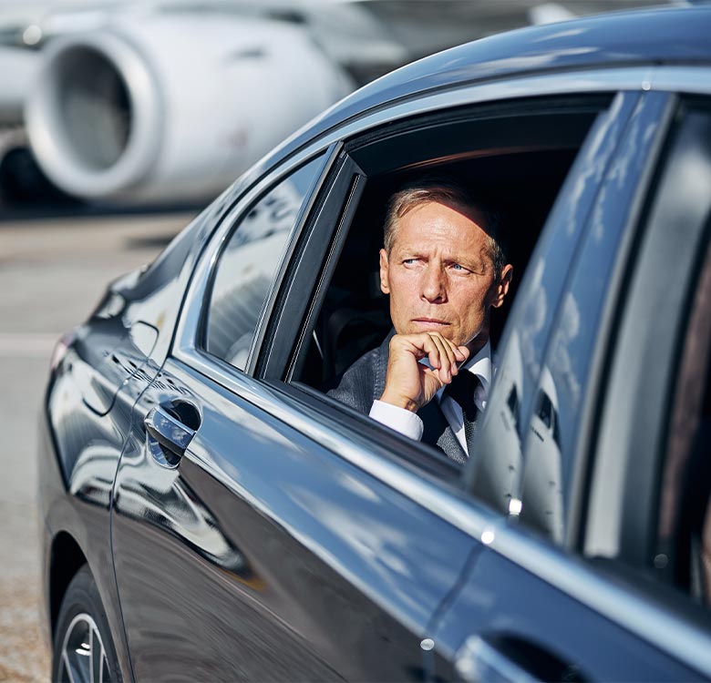 Businessman in a suit sitting inside a luxury car with a thoughtful expression, airport runway and airplane in the background, symbolizing travel and executive transportation.