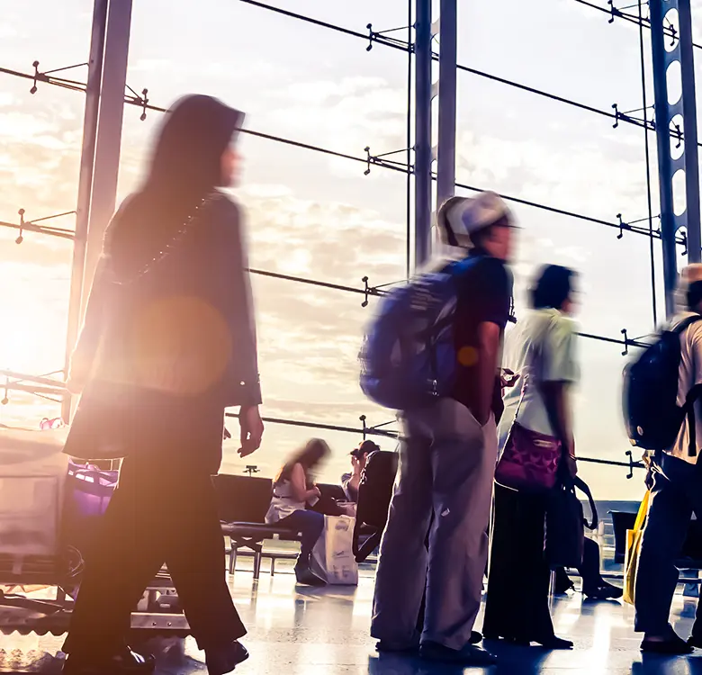 Crowded airport terminal with travelers moving through a modern space featuring large glass windows and natural light, showcasing a busy travel scene.