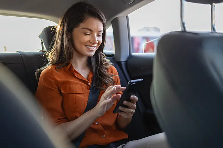 Young woman sitting in the backseat of a car, smiling while using her smartphone, dressed in a casual orange shirt, with sunlight illuminating the interior.
