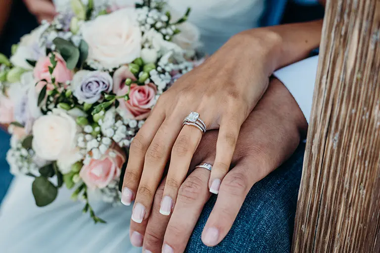 Close-up of a bride and groom's hands, showcasing elegant wedding rings. The bride's hand holds a bouquet of pastel roses and greenery, while the groom's hand rests on hers, symbolizing love and commitment. The background features a rustic setting, enhancing the romantic atmosphere.