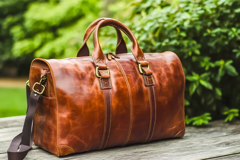 Brown leather duffel bag with sturdy handles and an adjustable shoulder strap, resting on a wooden surface in a lush outdoor setting.