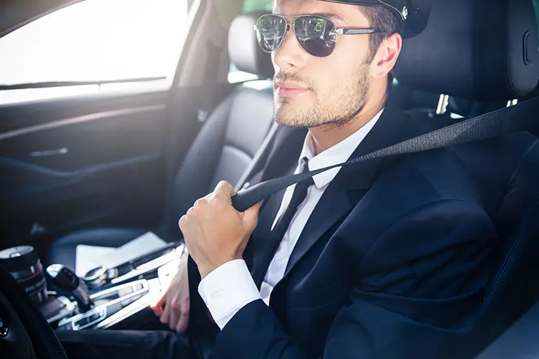 Professional chauffeur wearing sunglasses and a suit, seated in a luxury car with a seatbelt fastened, ready to drive passengers.