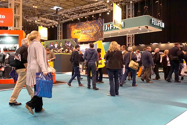 Crowd of attendees at a photography trade show navigating the exhibition floor, with prominent brand booths like Nikon and Fujifilm in the background. The scene captures the vibrant atmosphere of industry professionals and enthusiasts engaging with the latest photography technology.