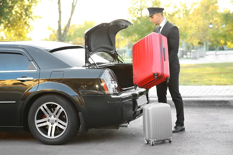 Professional chauffeur loading bright red suitcase and gray luggage into the trunk of a black luxury car, set against a backdrop of a sunny outdoor scene with trees.