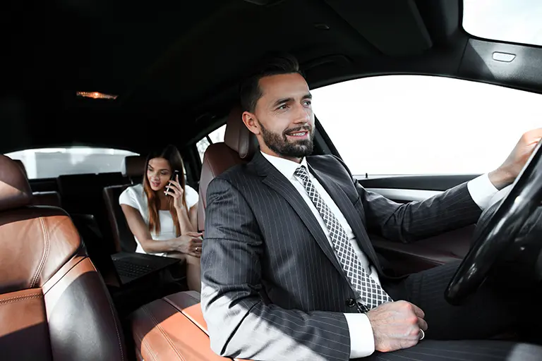 Professional male driver in a suit driving a luxury car, while a woman in a white dress sits in the back seat talking on the phone. The interior features modern design with brown leather seats.