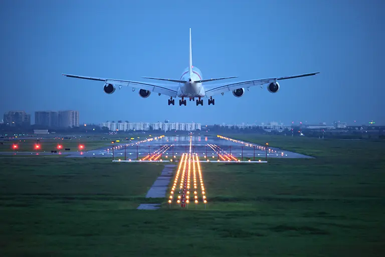 Airplane landing at dusk on a well-lit runway, showcasing airport infrastructure and city skyline in the background.